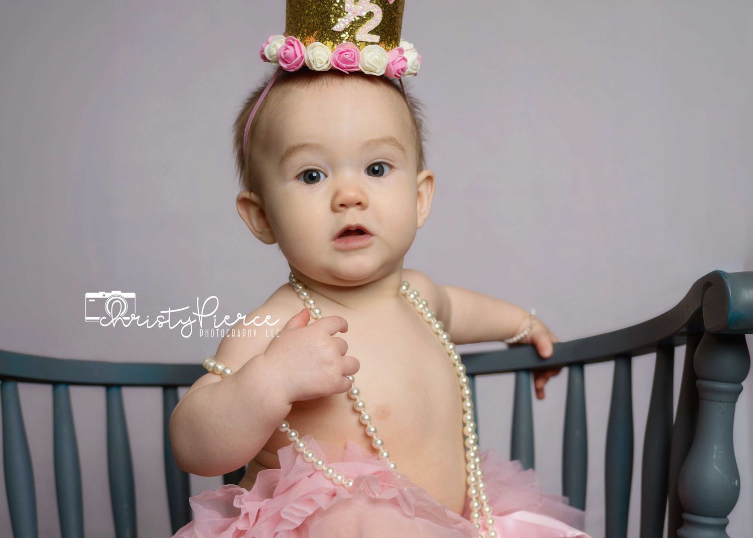 Baby in pink tulle dress and gold crown sits in dark crib for birthday photoshoot.
