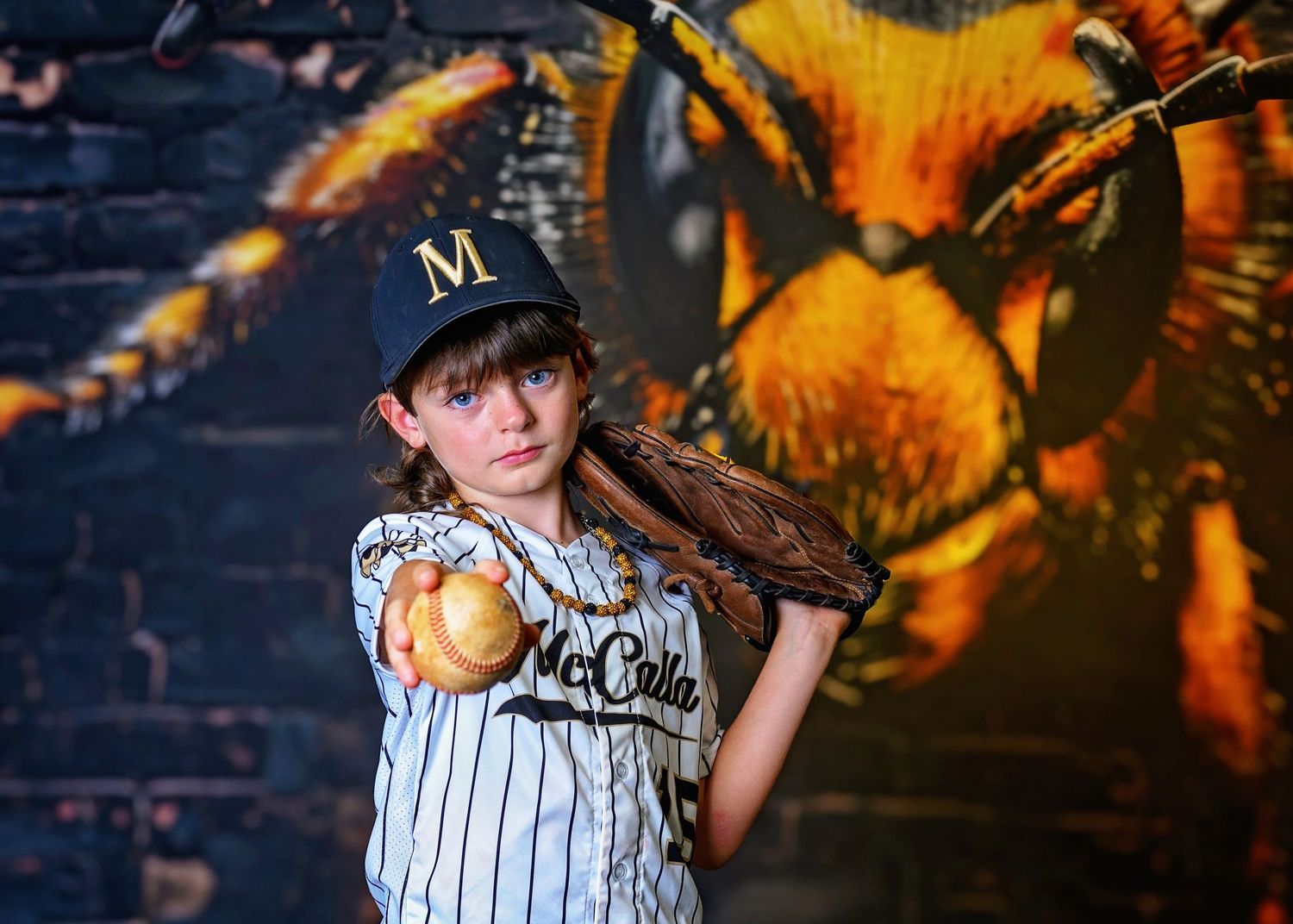 Youth baseball player in pinstripe uniform poses with bat against dramatic orange and black graffiti wall background.