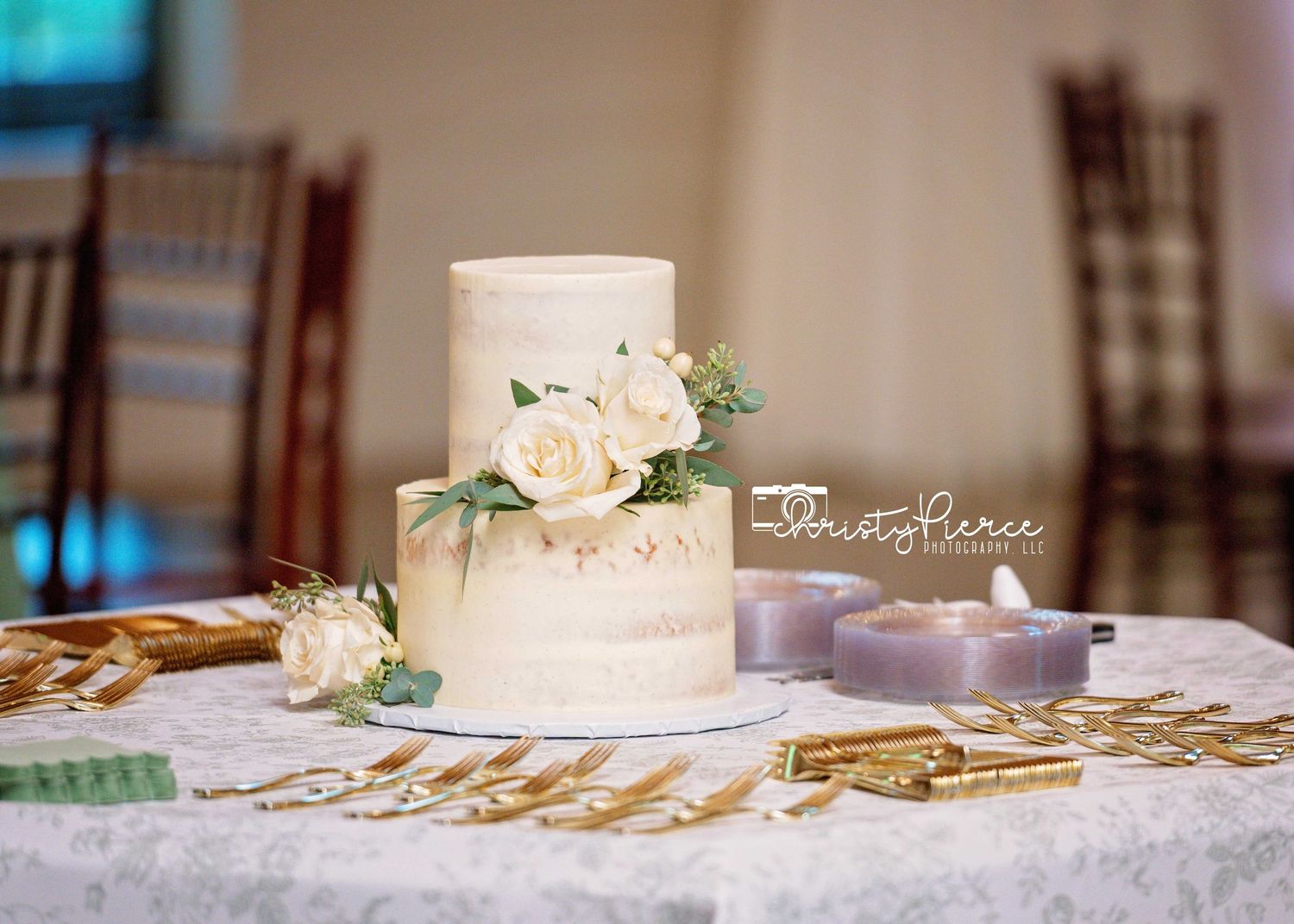 Elegant white wedding cake with fresh roses and greenery on a lace tablecloth with gold accents and purple candles.
