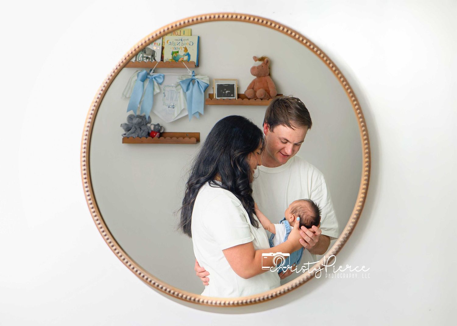 Parents tenderly hold their newborn baby inside a round wooden frame, with nursery decor on a shelf behind them.