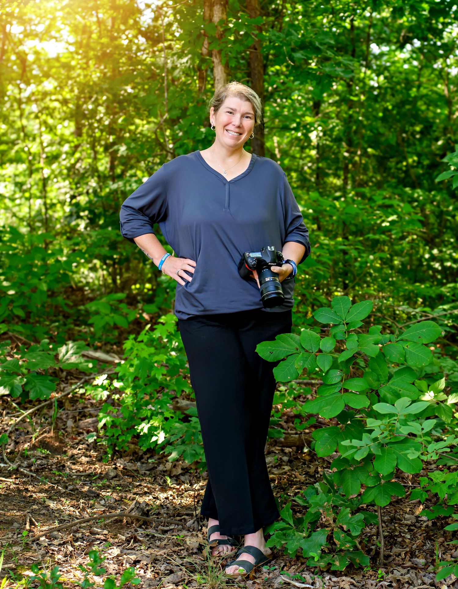 Smiling female photographer holding a DSLR camera standing in a lush green forest wearing a blue top.