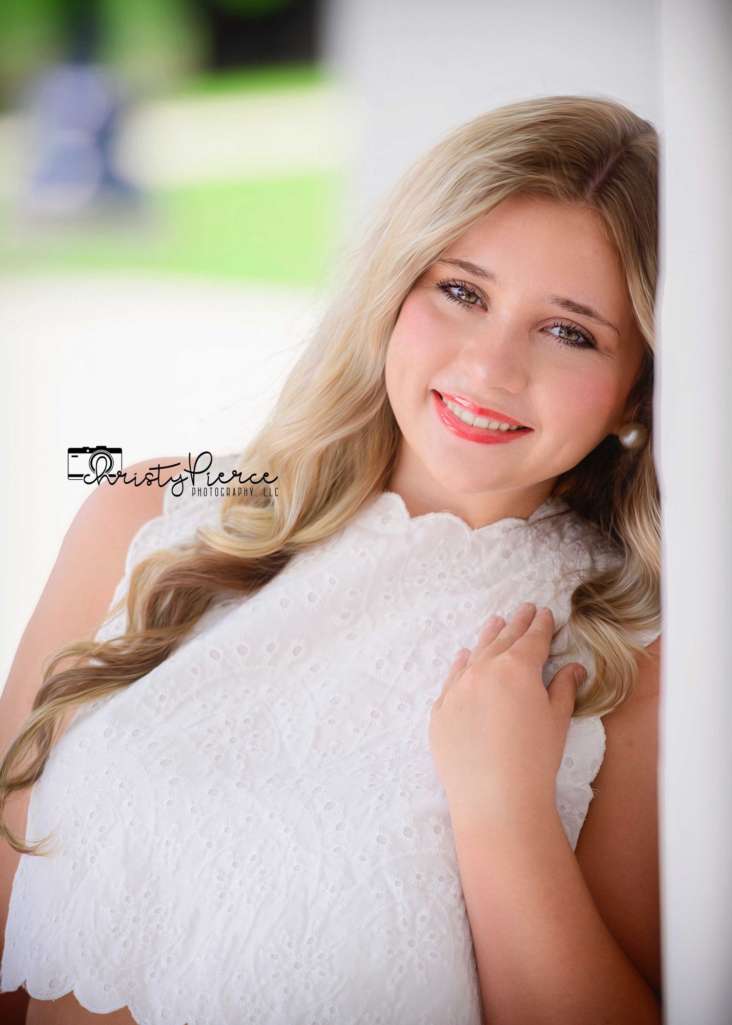 Young blonde woman in white lace dress smiling warmly, leaning against a white pillar outdoors.