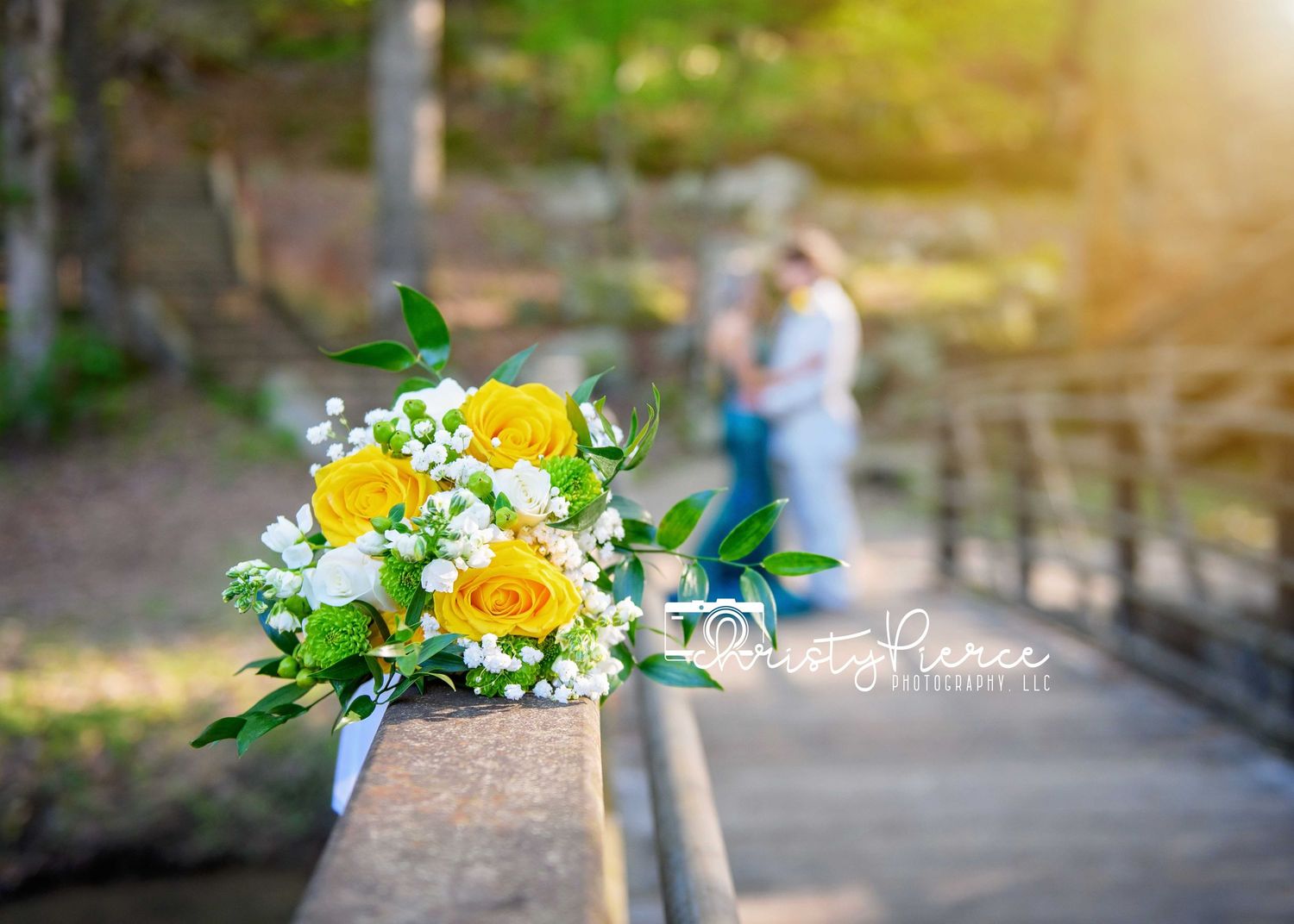 Yellow rose wedding bouquet on wooden railing with blurred couple embracing in background at sunset.