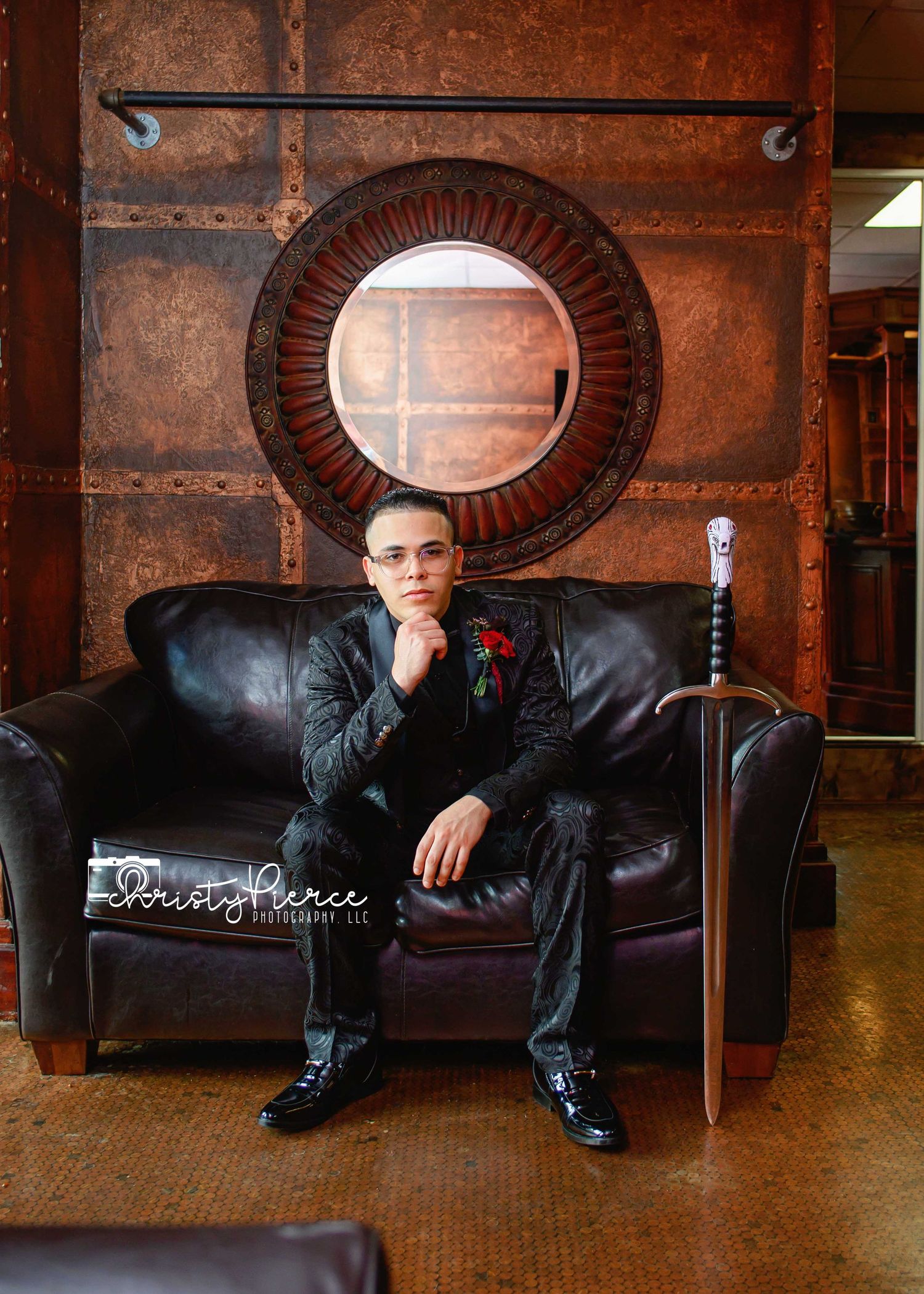 Young man in black suit with red boutonniere sits on leather couch beside a decorative sword in an elegant studio setting.