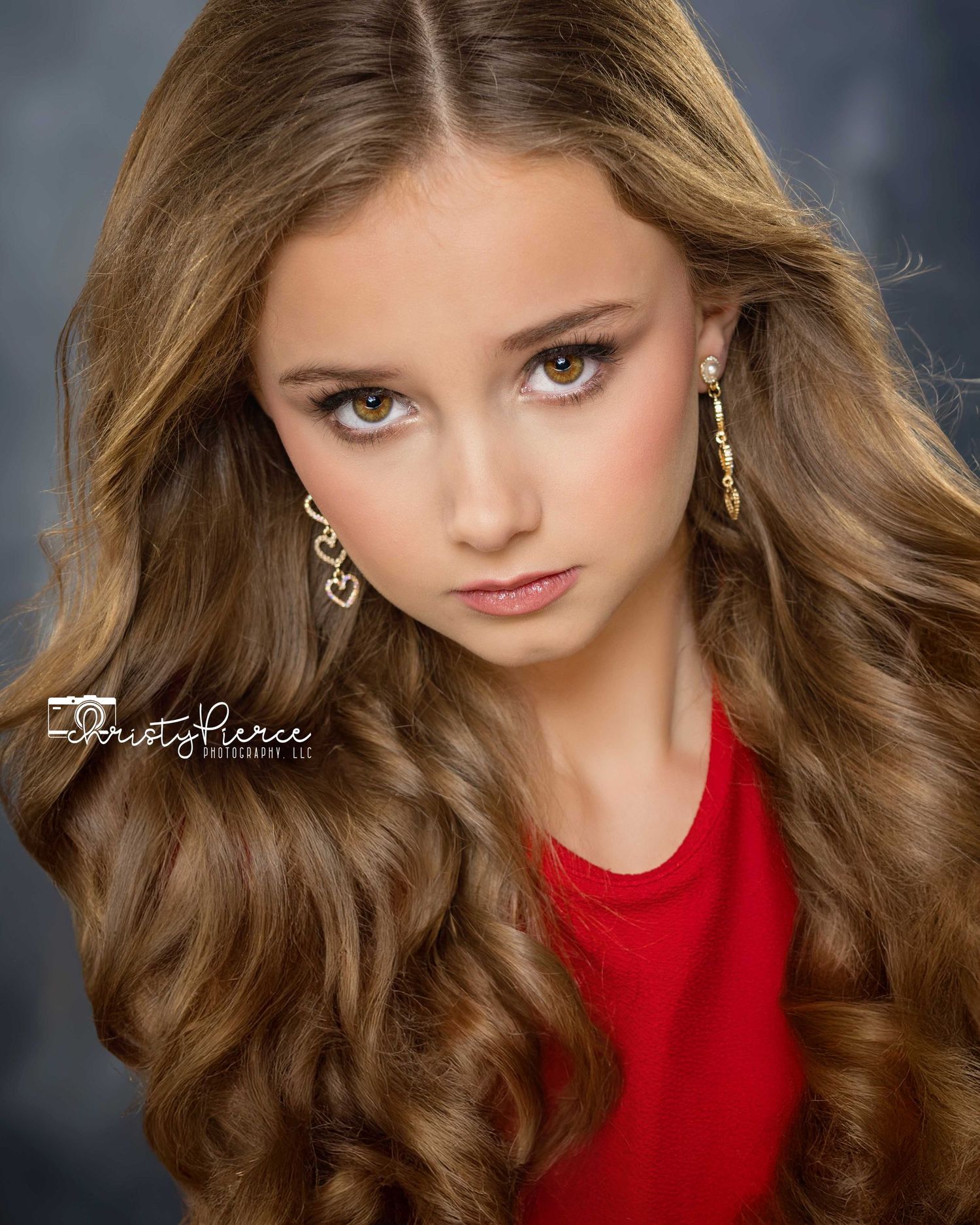 Professional portrait of a model in red wearing gold earrings with flowing wavy brown hair against dark background.