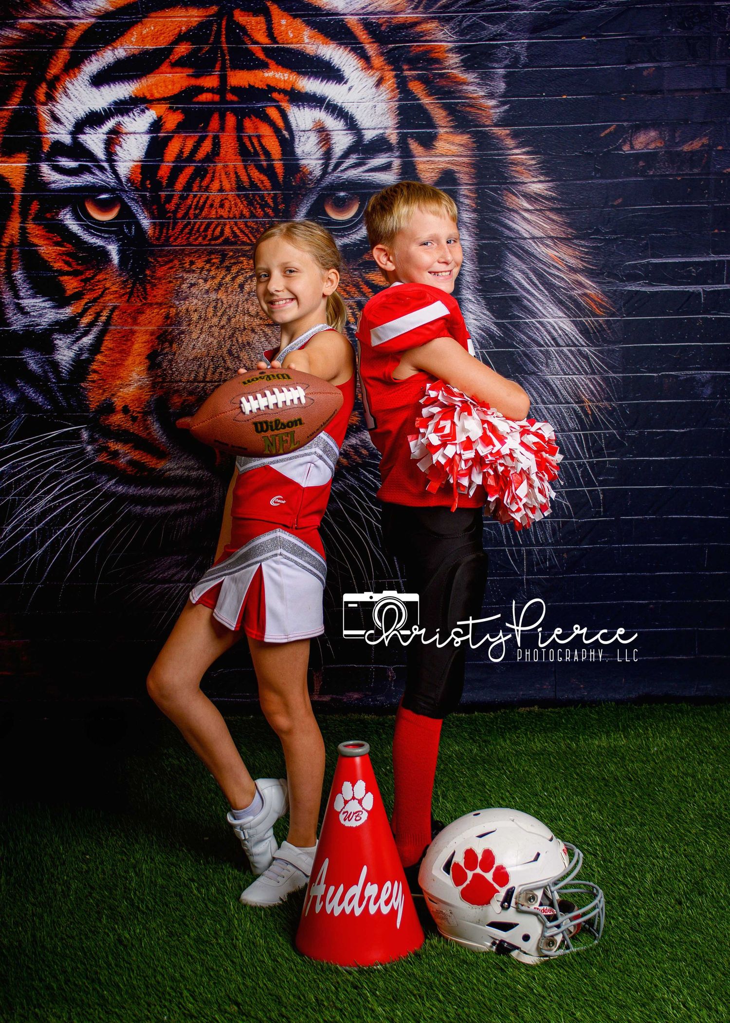 Two children in red cheerleader and football uniforms pose with a football and pom-poms in front of a tiger mural backdrop.