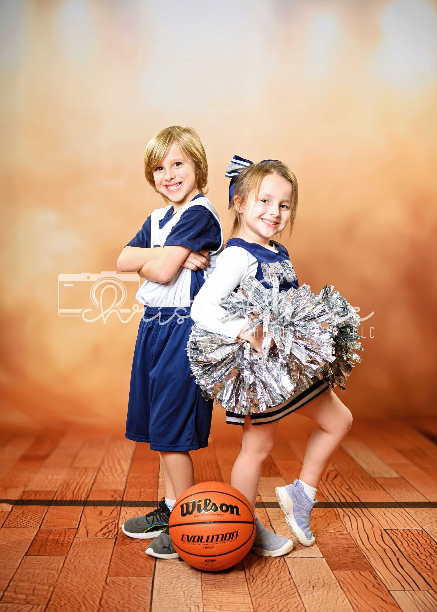 Two children in athletic attire pose on wooden floor with basketball and cheerleading pom poms.