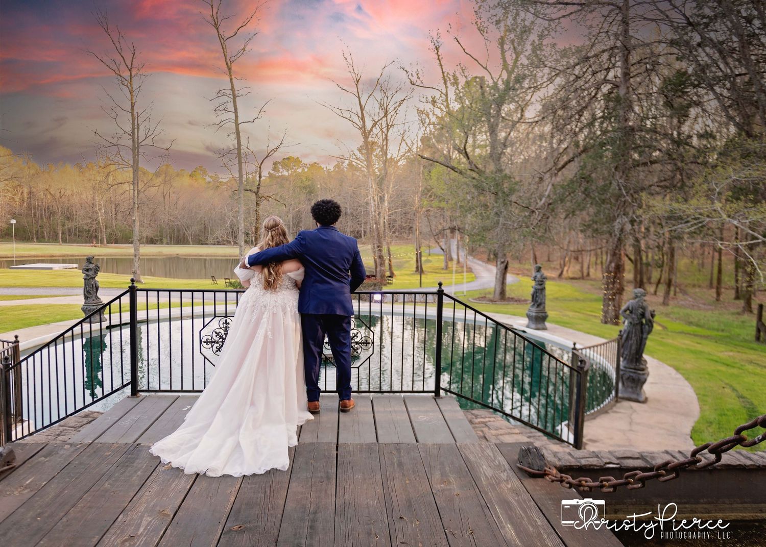 A couple in wedding attire stands on a wooden bridge overlooking a scenic park during a colorful sunset.