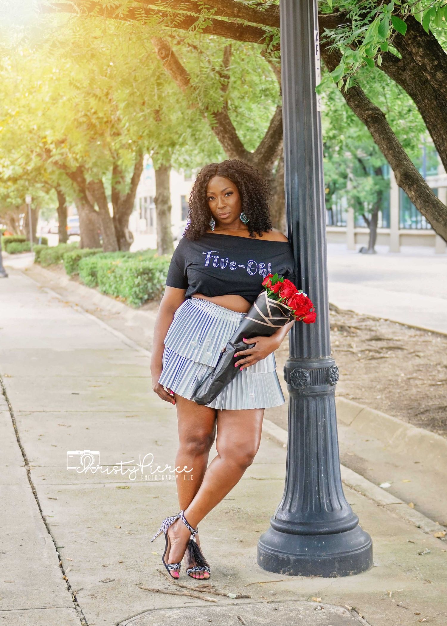 A person in a casual t-shirt and striped skirt poses next to a lamppost while holding red roses in a sunlit park setting.