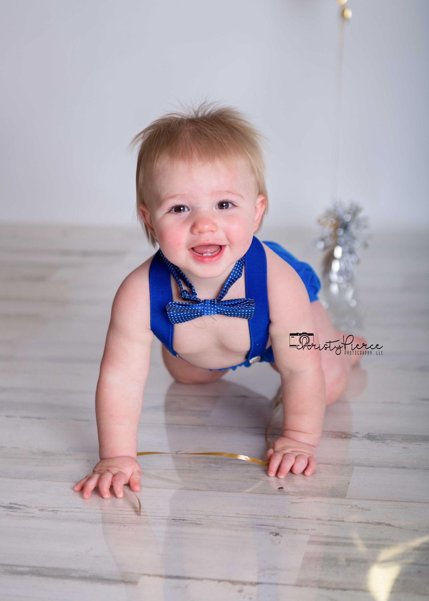 Smiling baby in royal blue bow tie and suspenders crawls on light wooden floor during professional photo session.