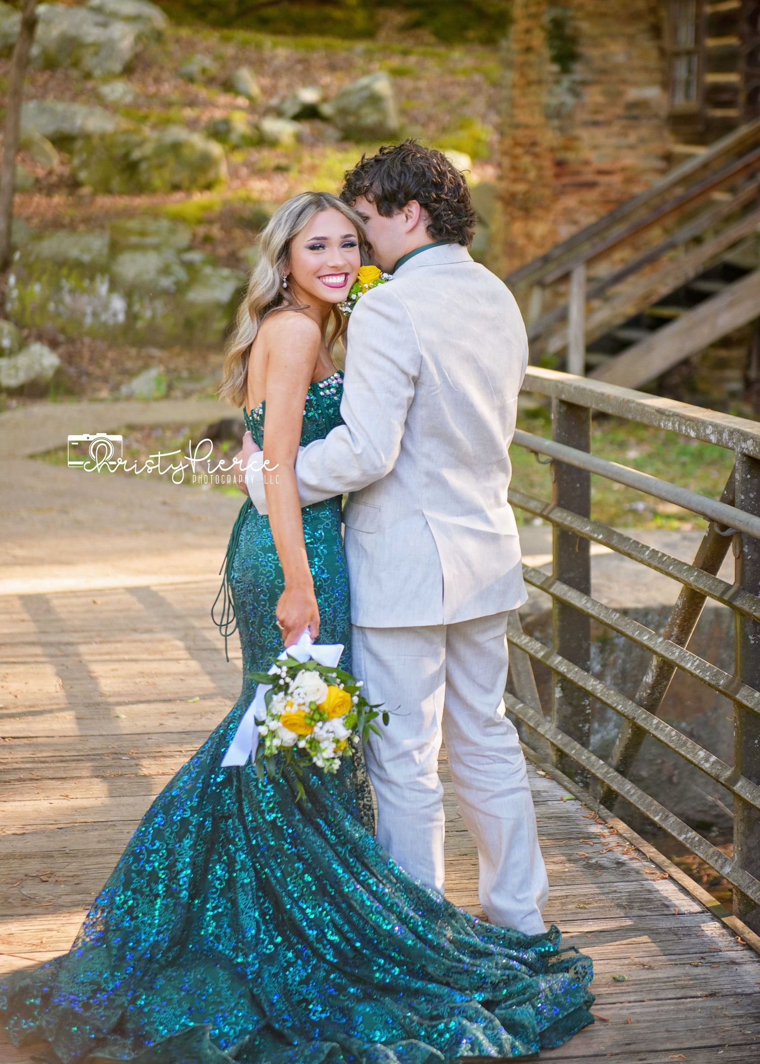 A couple shares a romantic moment on a wooden bridge while wearing a teal sequined gown and light-colored suit.