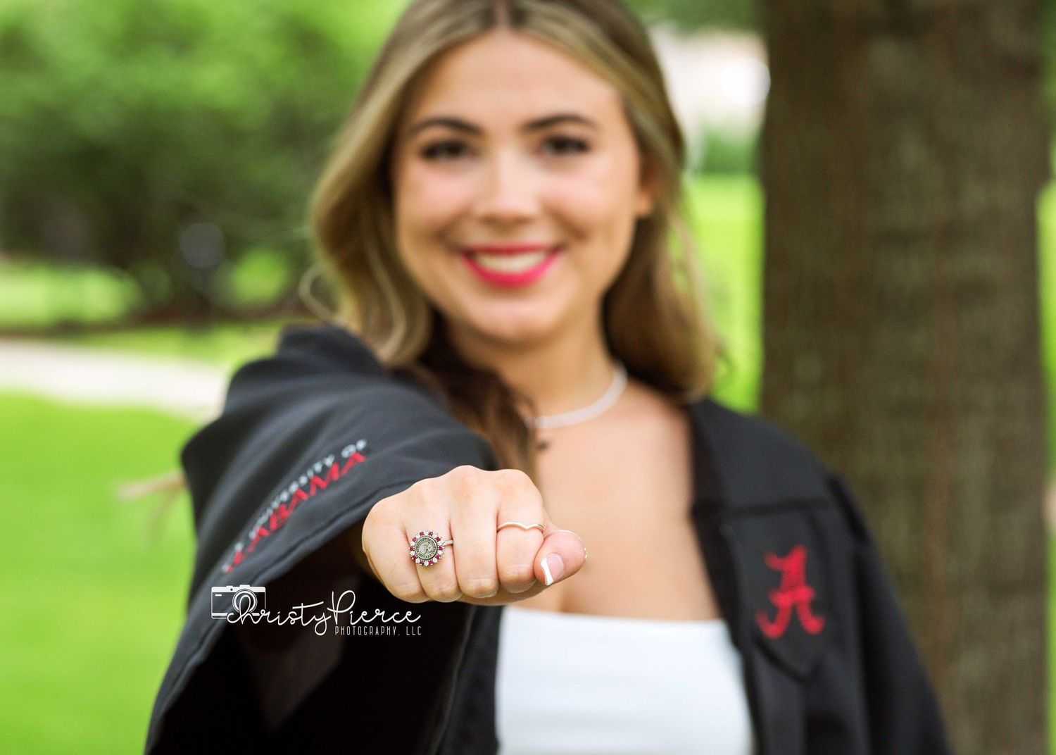 Graduate in white top and black graduation gown shows off class ring in outdoor portrait.