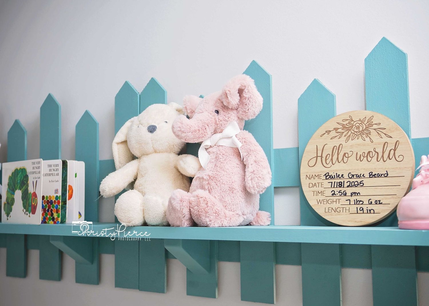 Plush toys and a decorative wooden sign displayed on a turquoise picket fence shelf against a white wall.