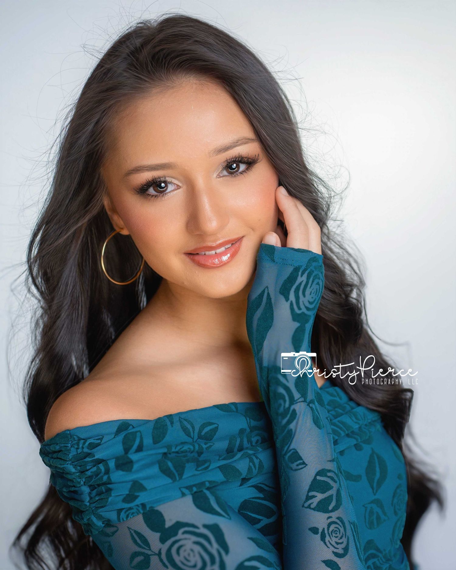 Young woman in teal off-shoulder floral top with gold hoop earrings, posing for a professional portrait photo.