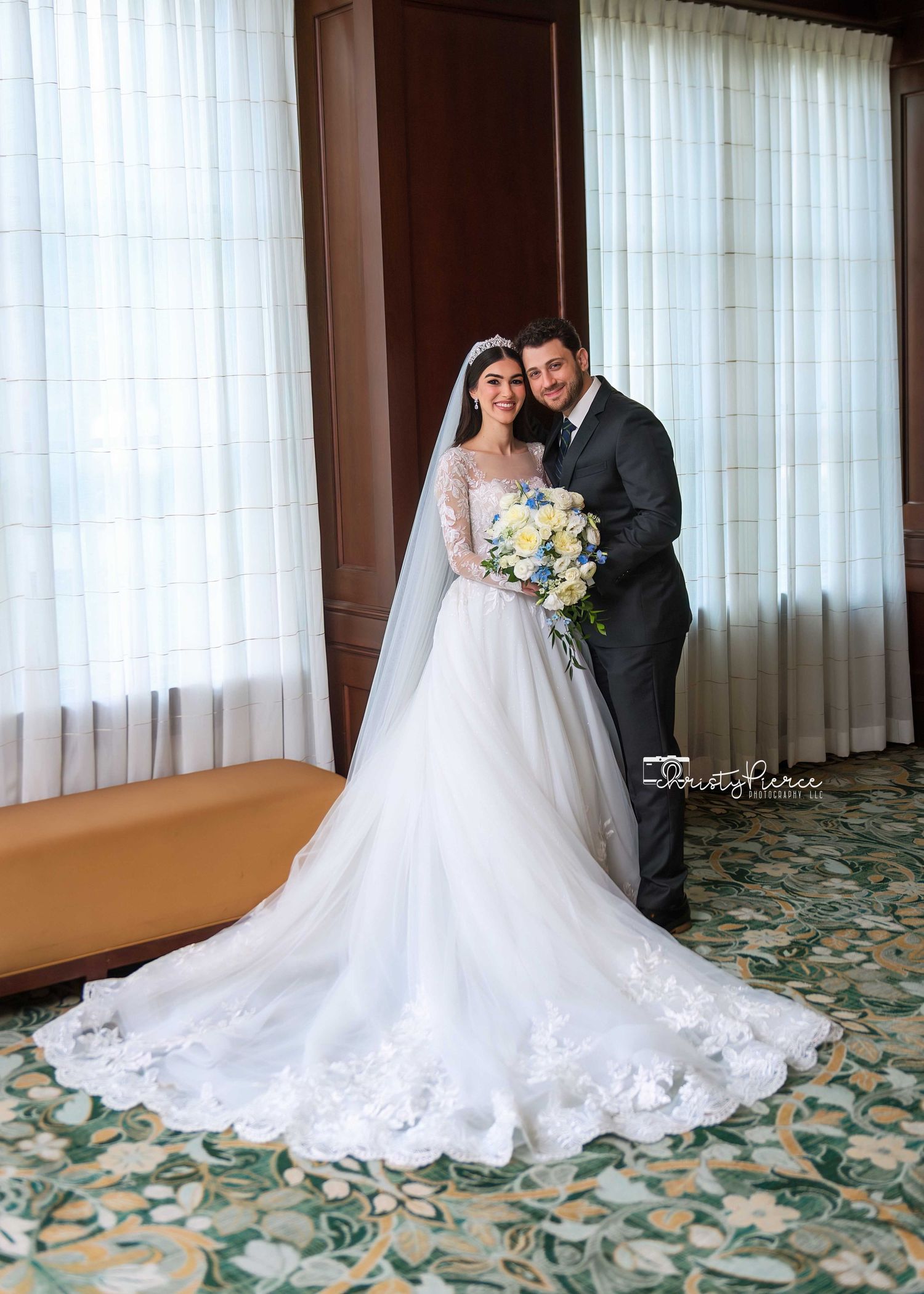 A bride and groom share an intimate moment in a hotel room with flowing white curtains and patterned carpet.
