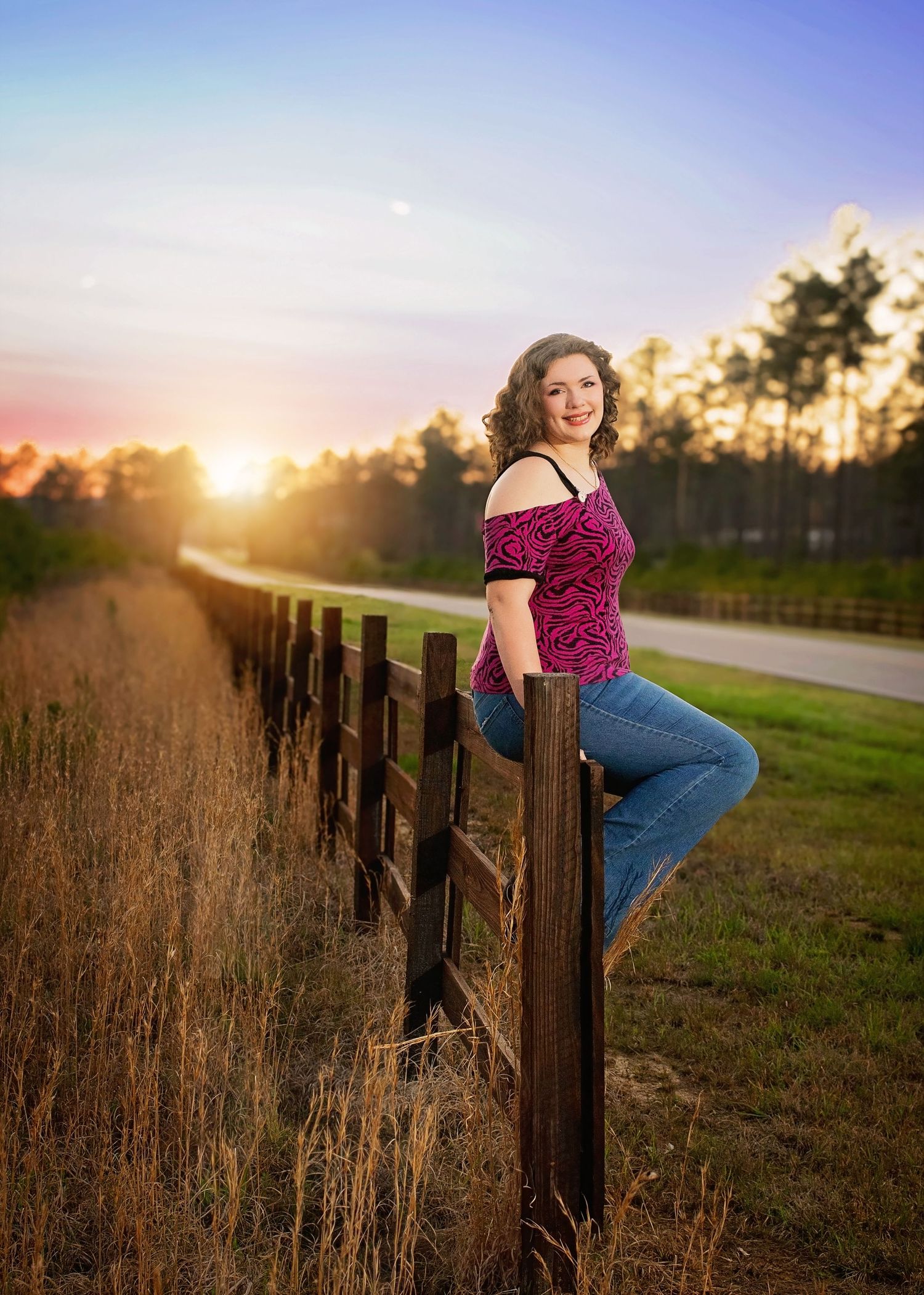 Young woman in pink top and jeans poses on wooden fence at golden sunset with tree-lined road backdrop.