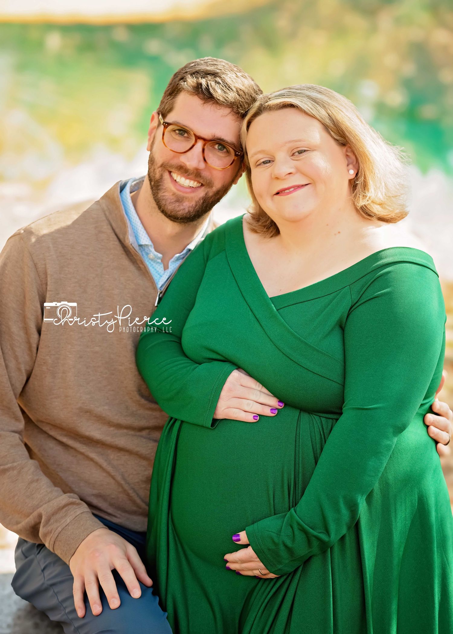 Couple in casual attire embracing outdoors during a maternity photoshoot with a green and natural background.