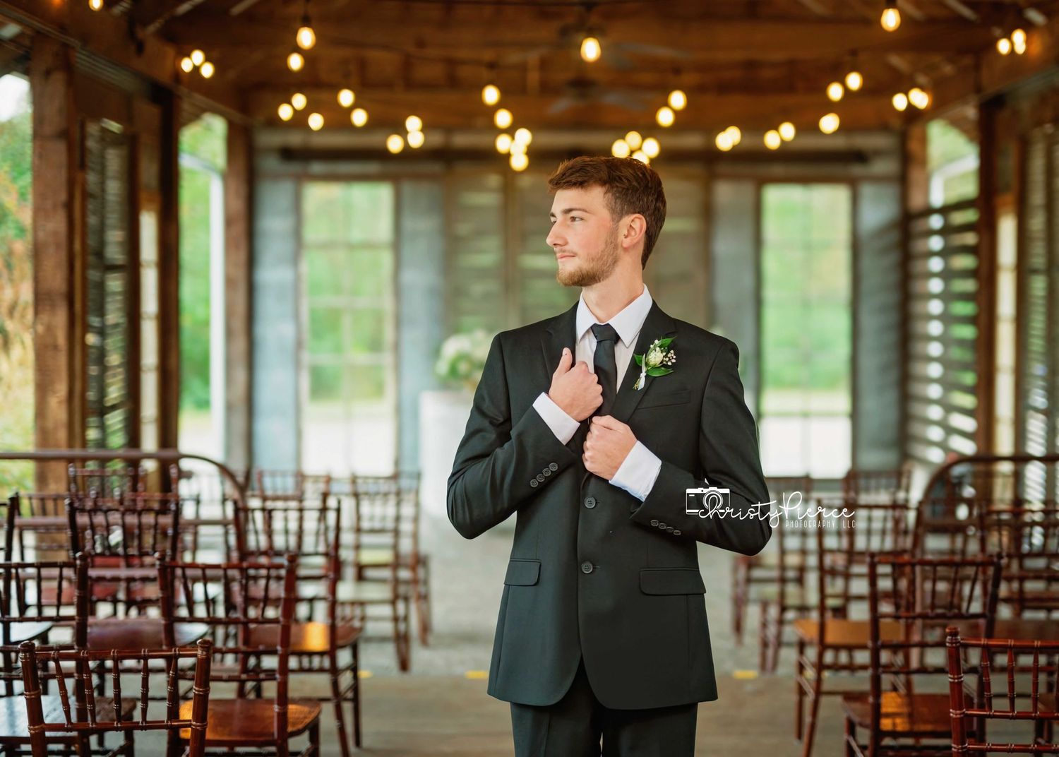 A person in a dark suit adjusts their tie while standing in an elegant reception venue with wooden chairs and warm lighting.