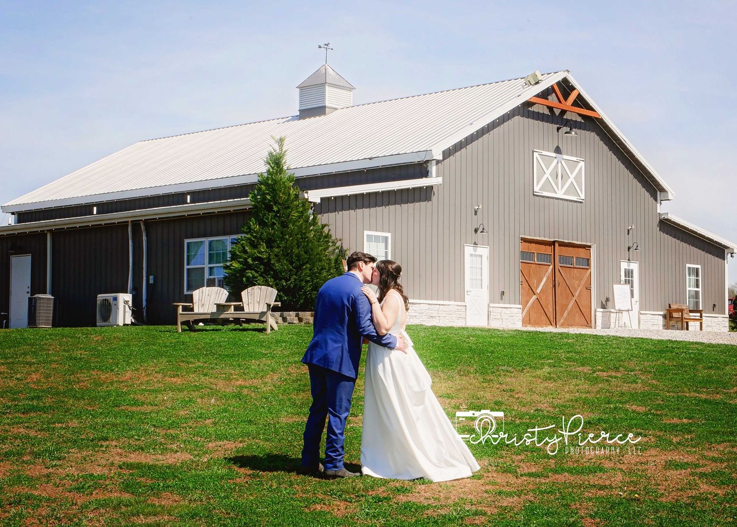 A bride and groom share a kiss on a green lawn in front of a rustic gray barn wedding venue on a sunny day.