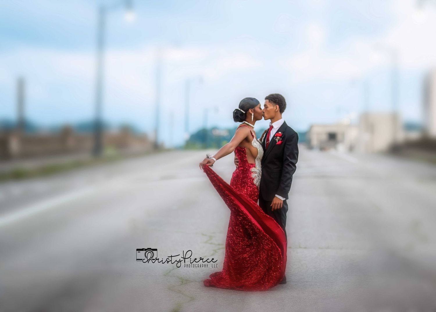 A couple in formal attire poses romantically on an empty road with a flowing red dress creating dramatic movement.
