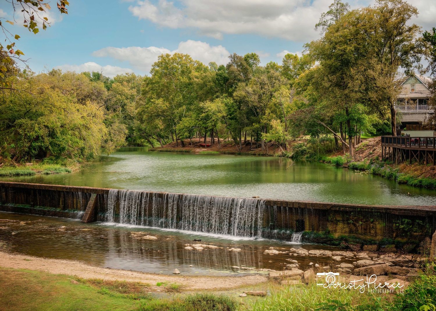 A scenic waterfall cascades over a stone dam surrounded by lush green trees and autumn foliage on a sunny day.