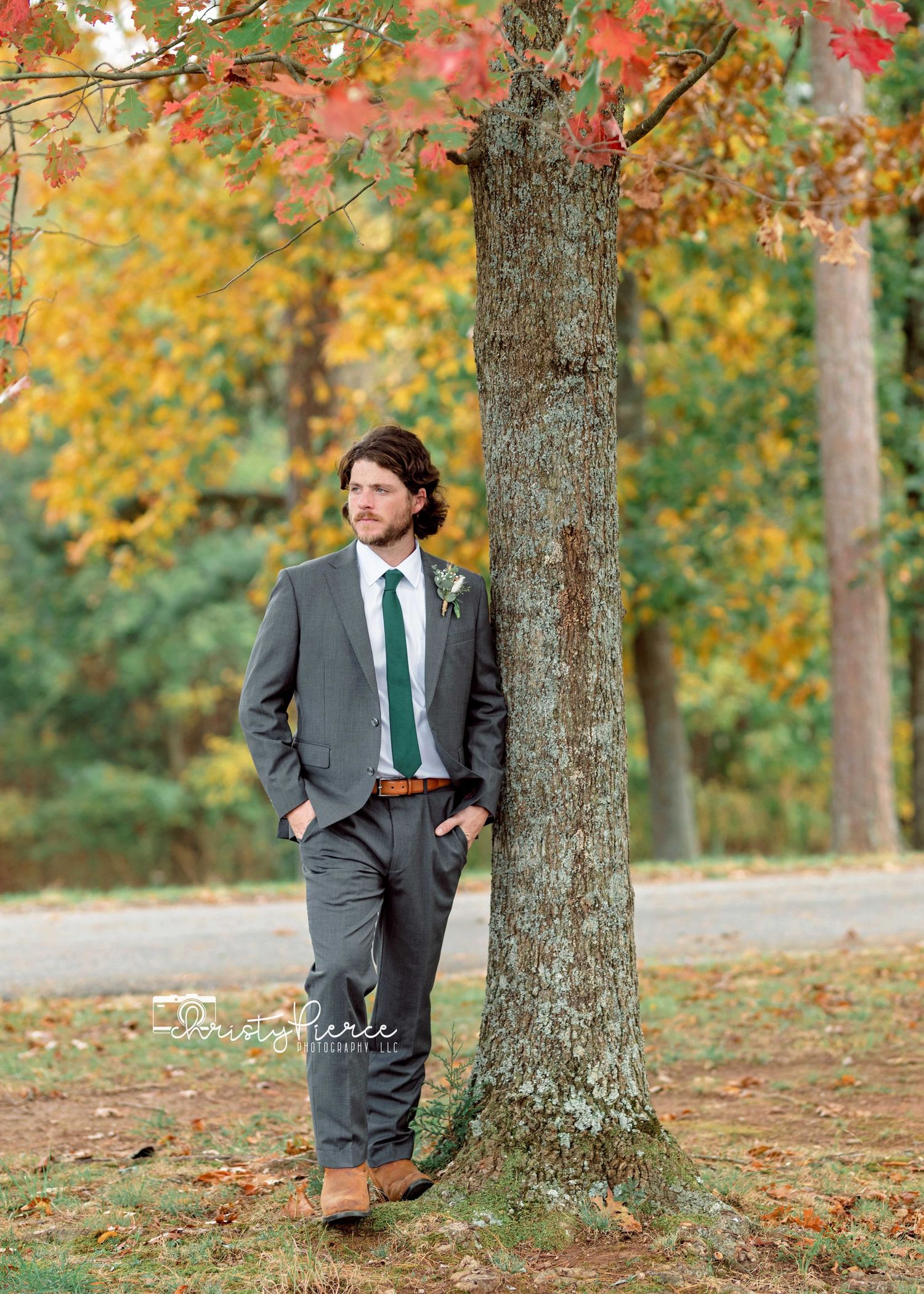 A person in a grey suit and green tie stands confidently next to a tree with vibrant fall foliage in the background.