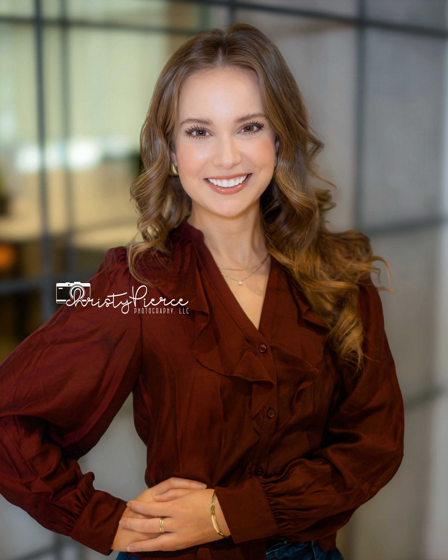 Professional portrait of someone wearing a burgundy button-up blouse with long wavy brown hair in an office setting.