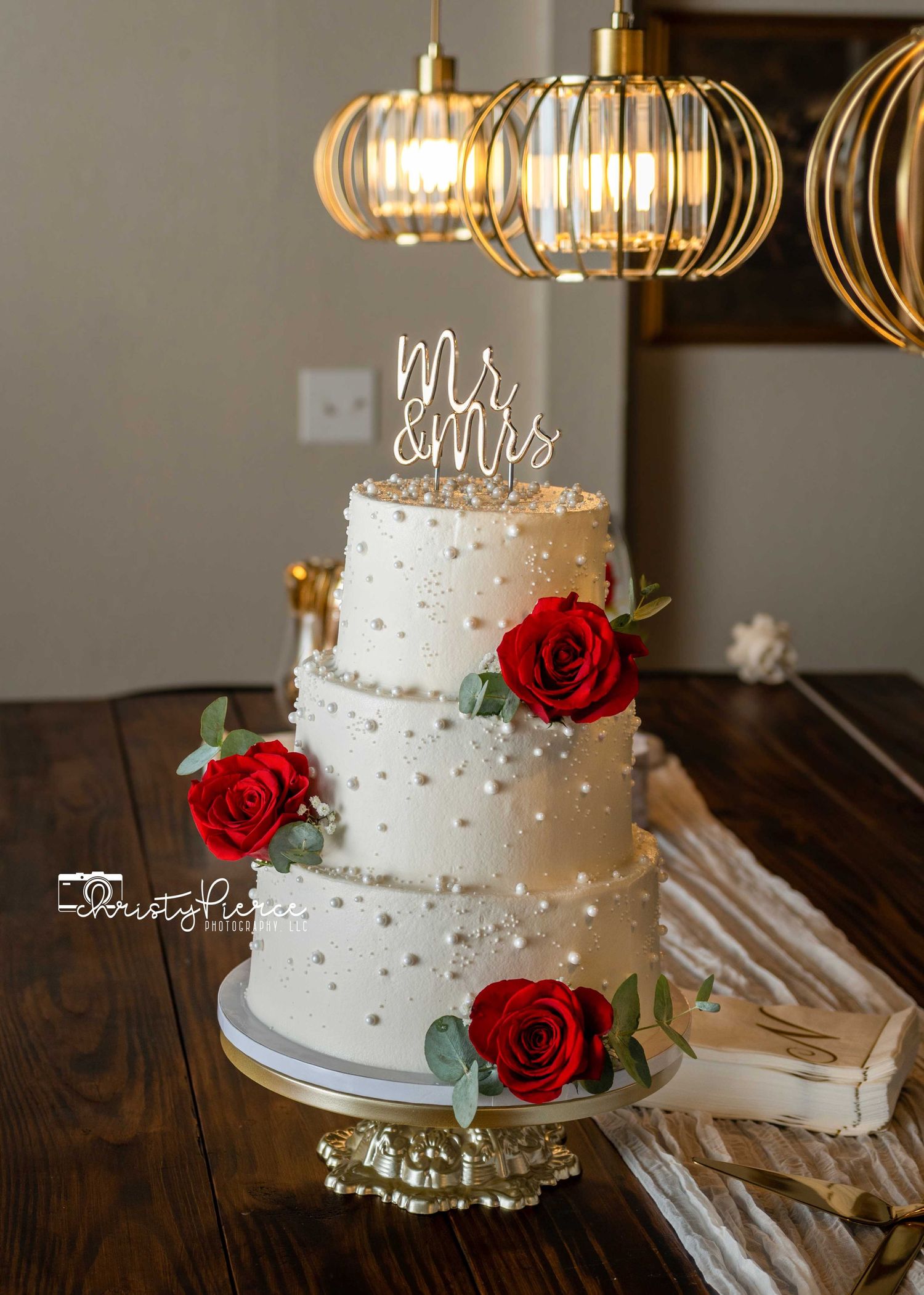 Three-tier white wedding cake with red roses and Mr & Mrs topper on a gold stand under pendant lights.