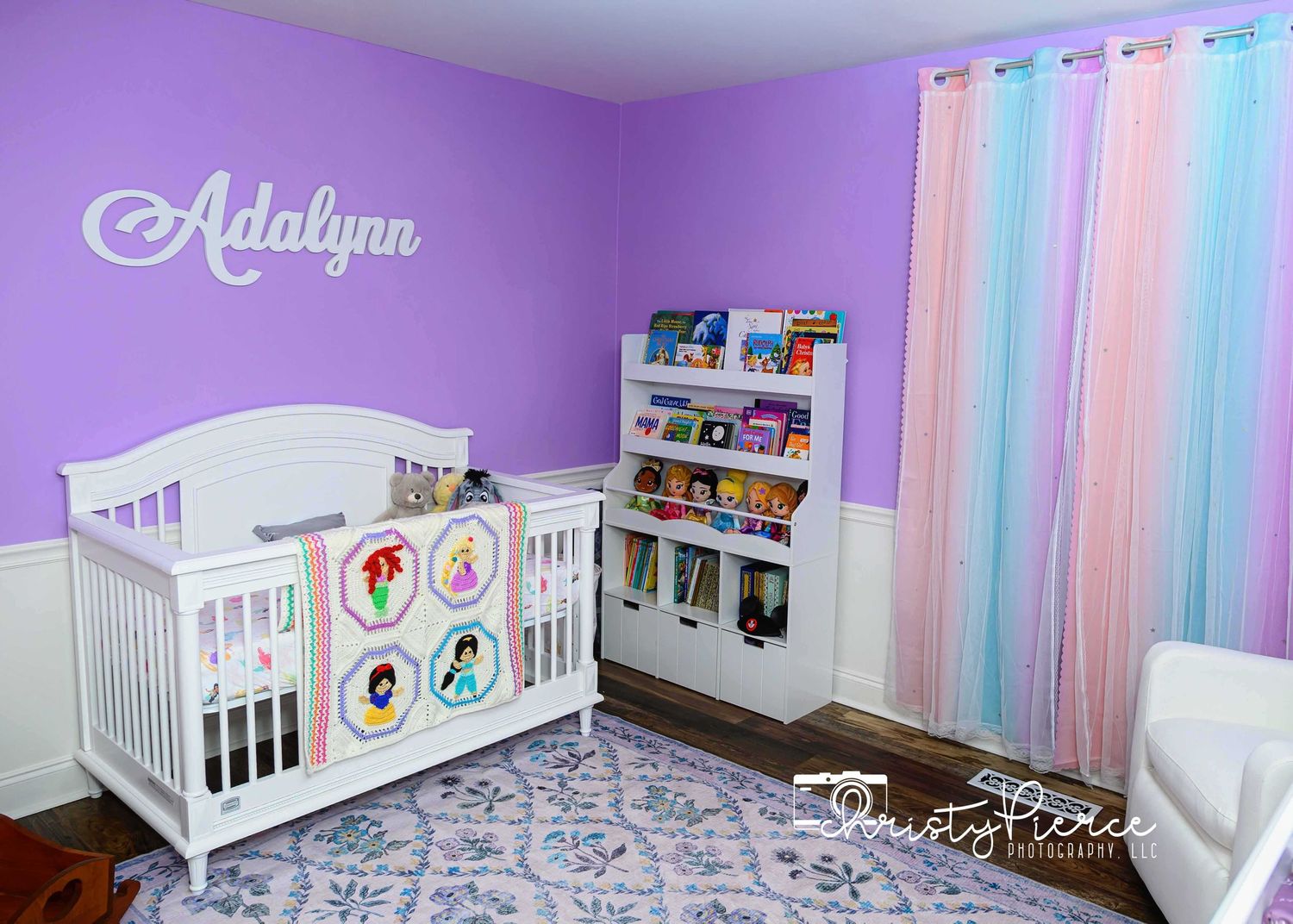 Purple nursery room with white crib, rainbow curtains, and book display shelf decorated with matching princess blanket.