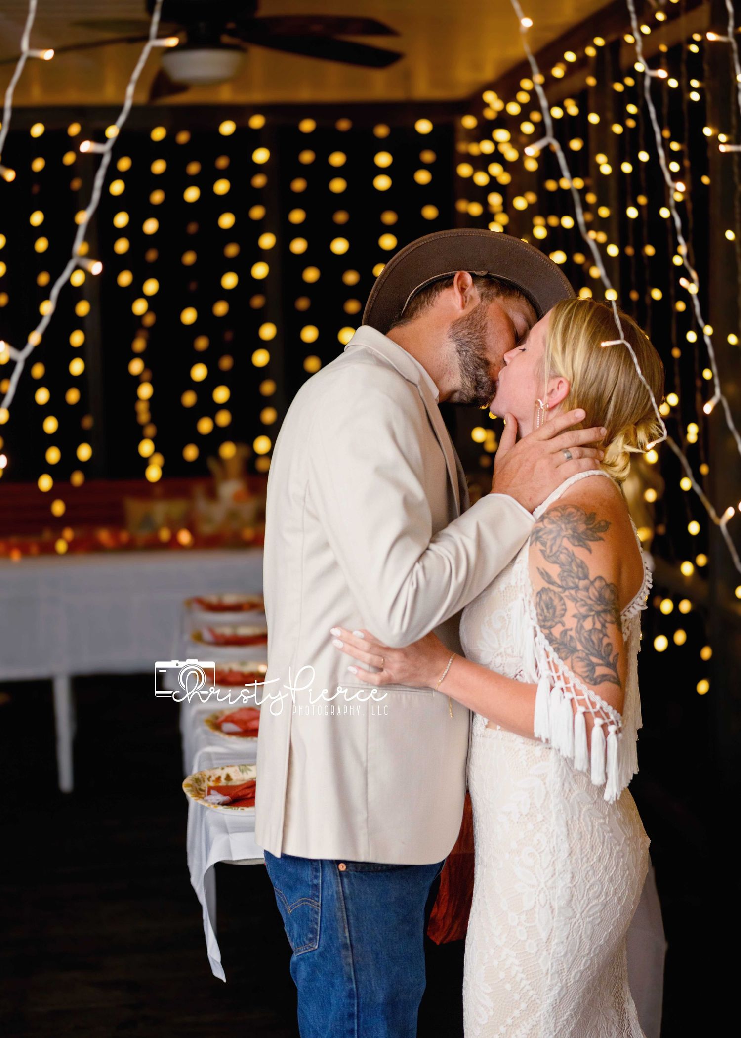 A couple shares a romantic kiss during their wedding celebration with twinkling lights in the background.
