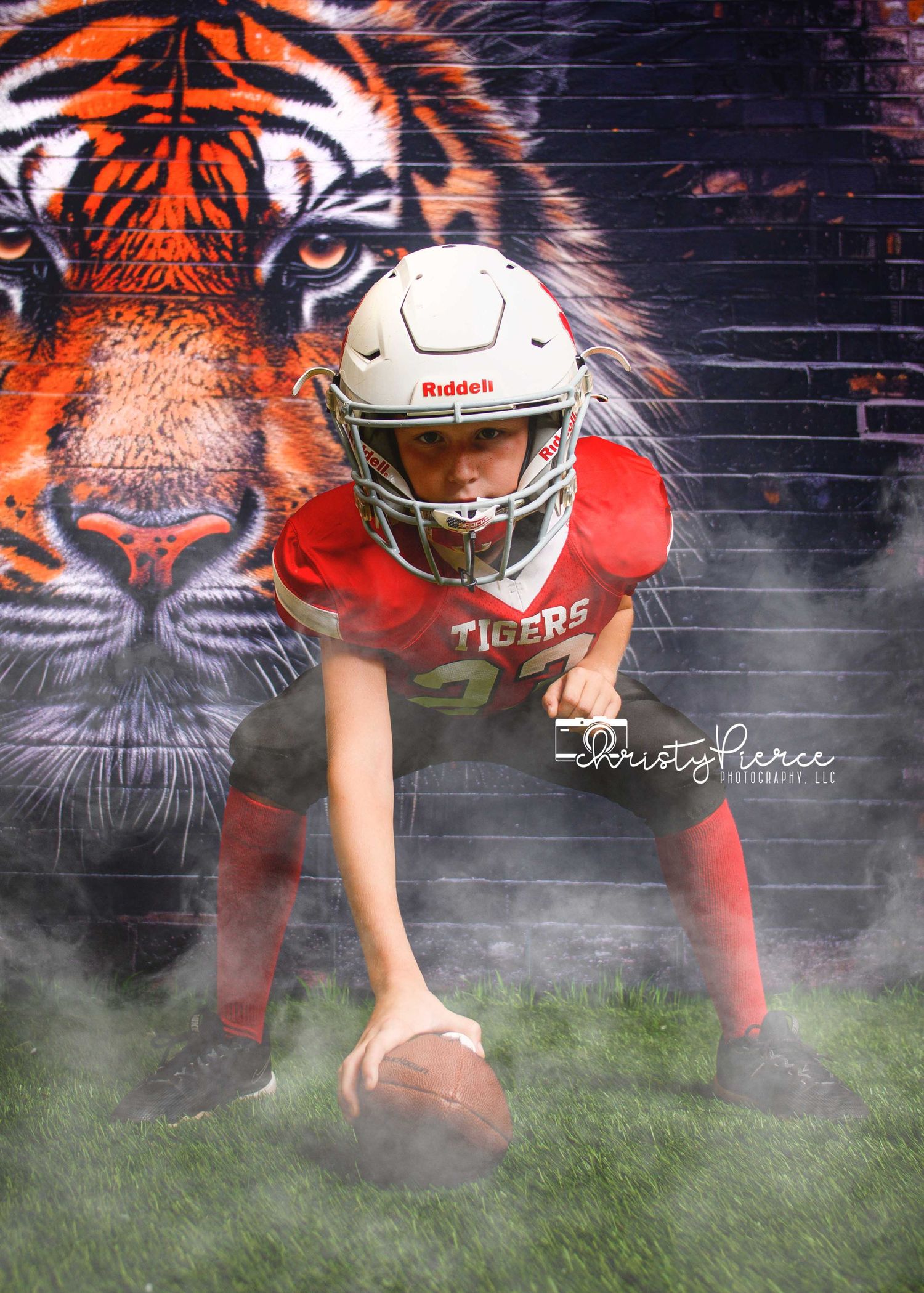 Young football player in red Tigers jersey and white Riddell helmet crouches over ball in front of tiger mural.