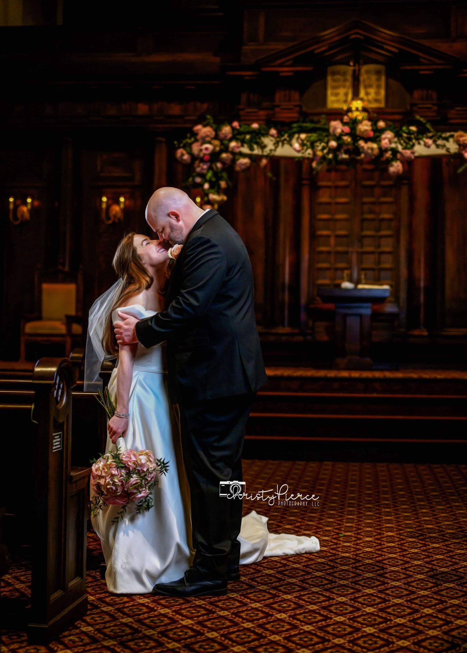 A romantic wedding kiss captured in a dimly lit church sanctuary decorated with floral arrangements.