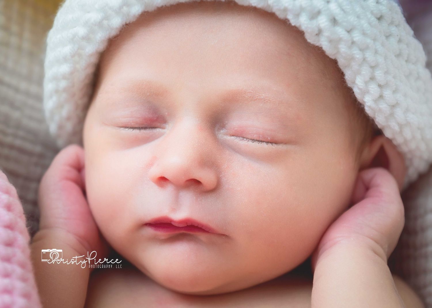 A sleeping newborn baby wearing a soft white knit hat rests peacefully against a gentle pink background.