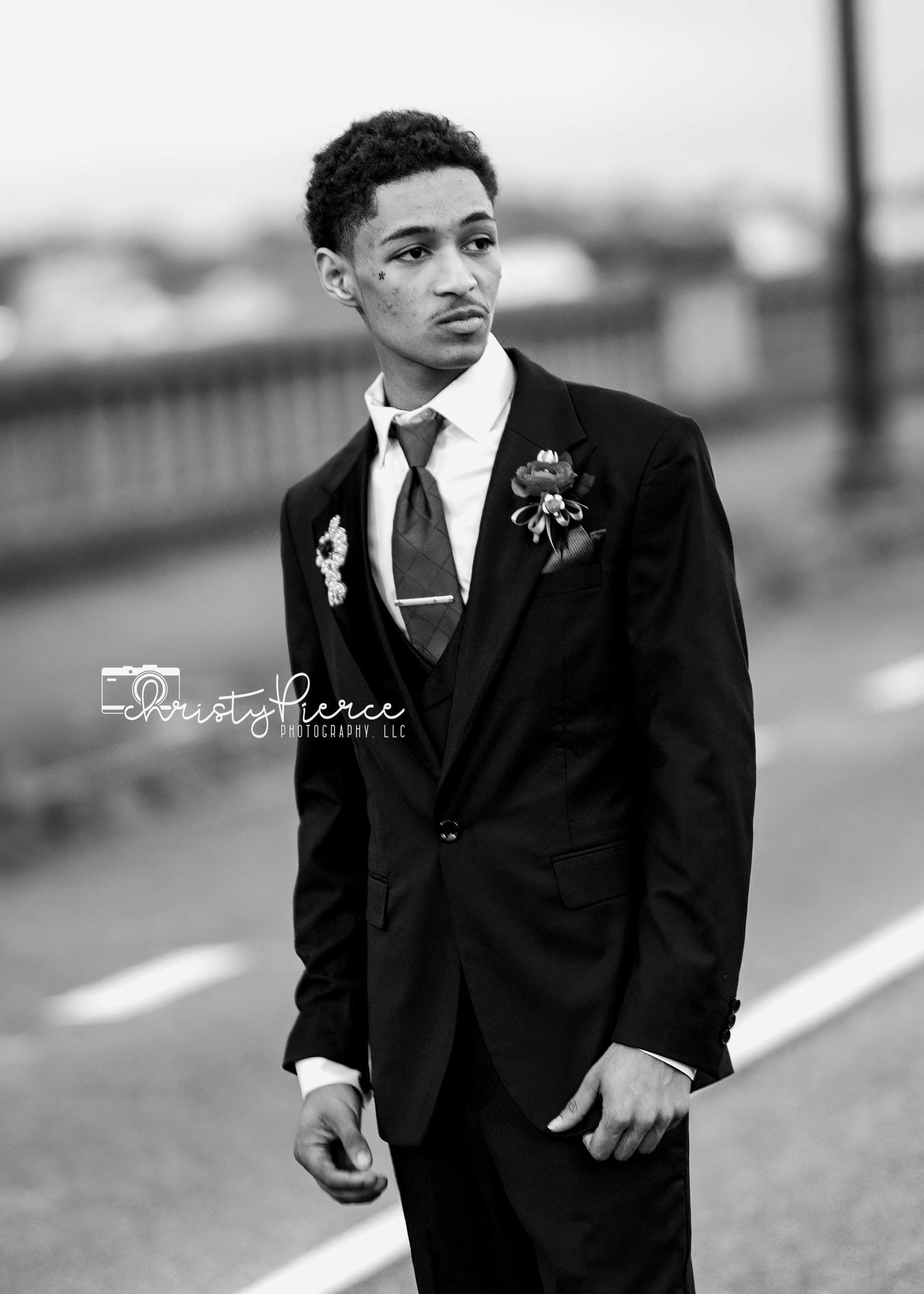 A young man in a formal black suit and tie poses for a black and white portrait outdoors.