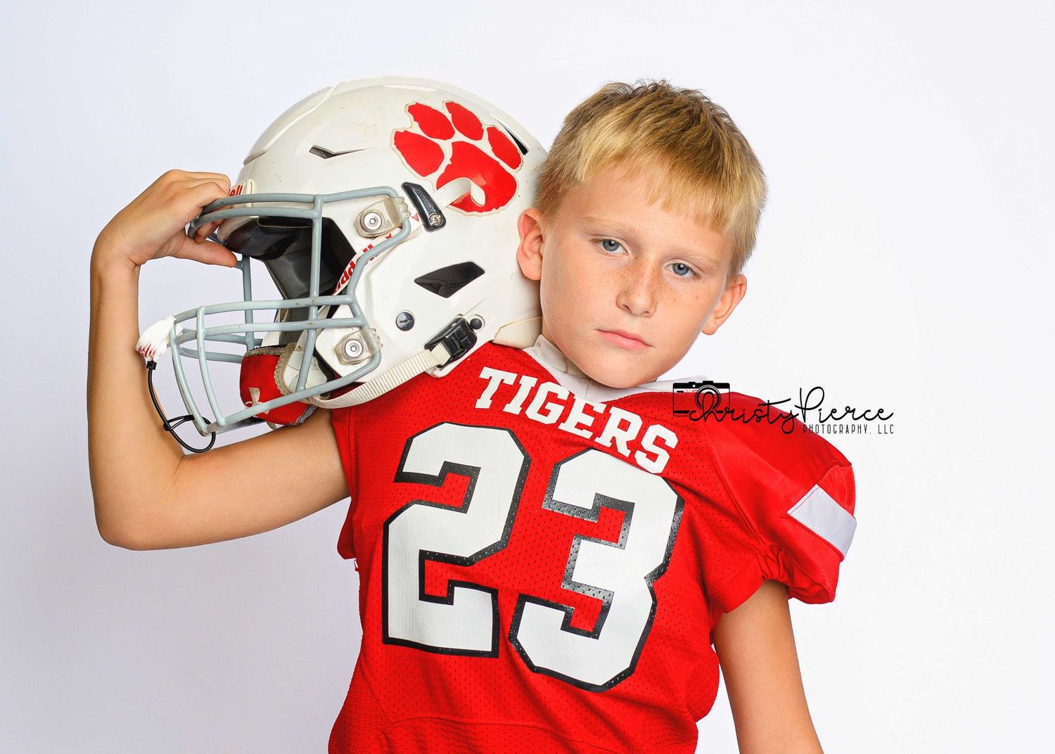 Young boy in red Tigers #23 football jersey poses with white helmet on shoulder against white background.