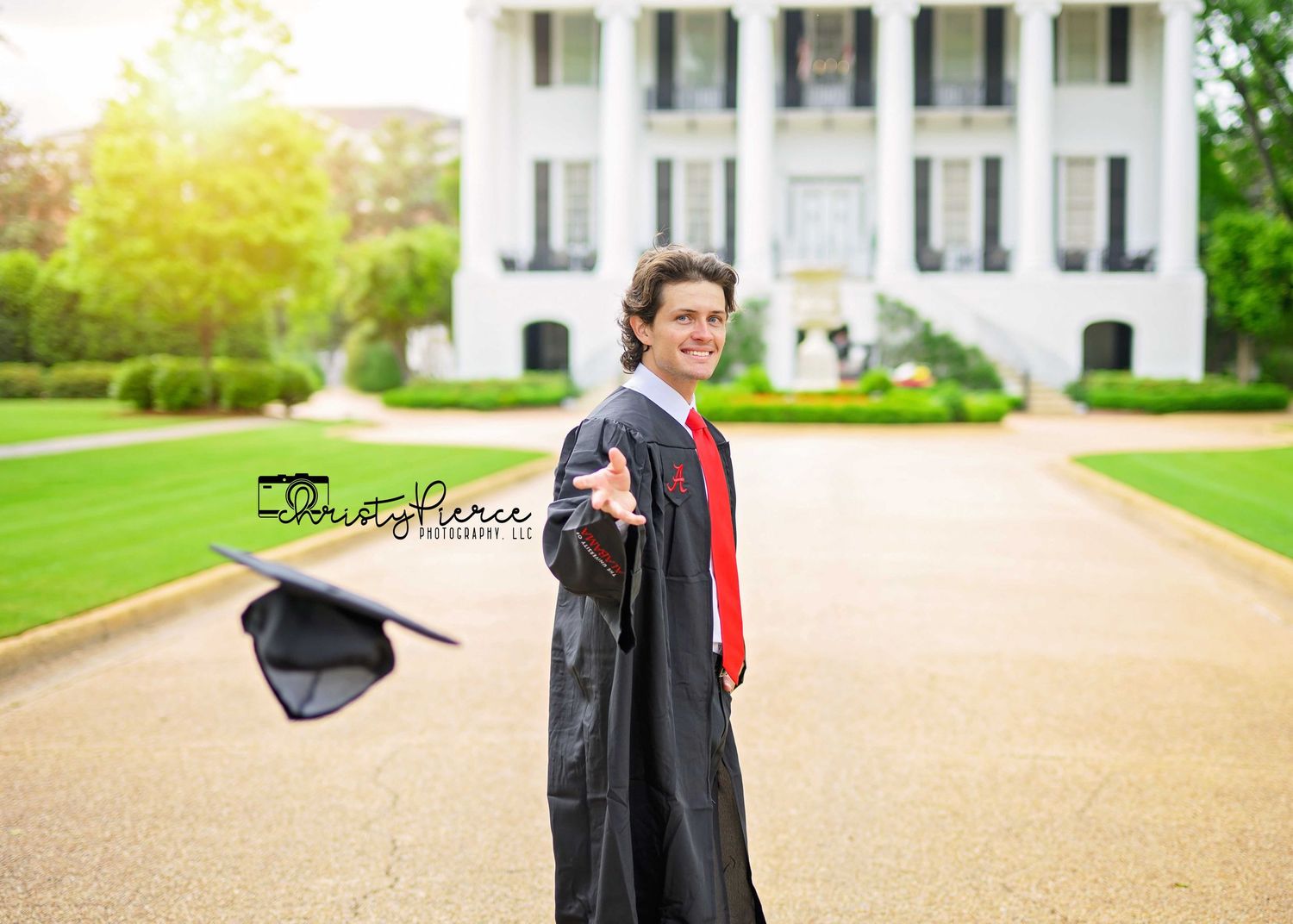Graduate in black cap and gown holds up diploma in front of stately white columned building on sunny day.