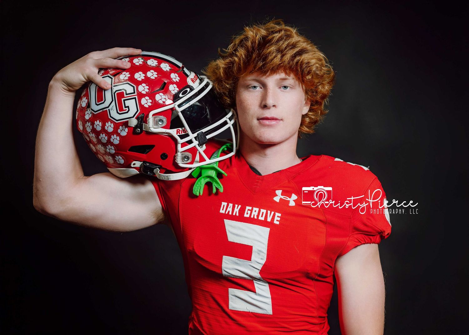 High school football player in red uniform poses with red and white patterned helmet against dark background.