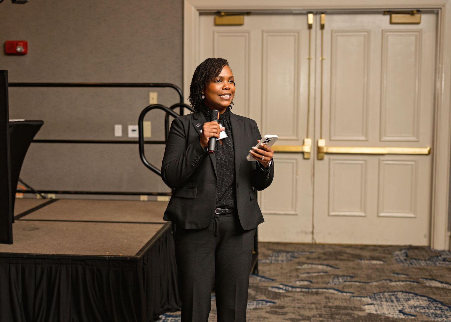 A speaker in a dark business suit holds a microphone while presenting on stage in a conference room setting.