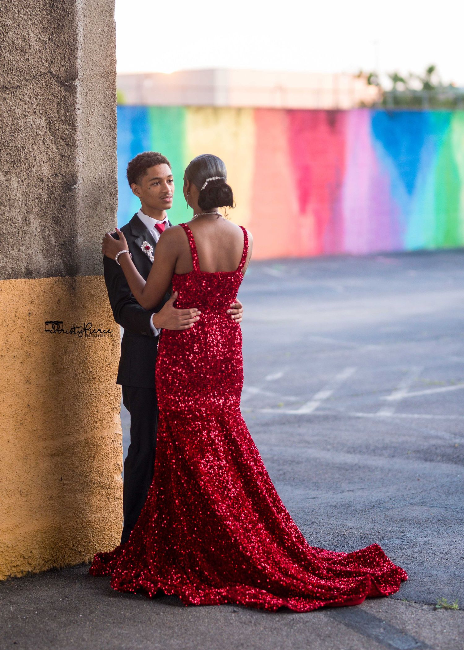 Couple in formal attire, with a stunning red sequined gown and dark suit, pose together against a colorful wall backdrop.