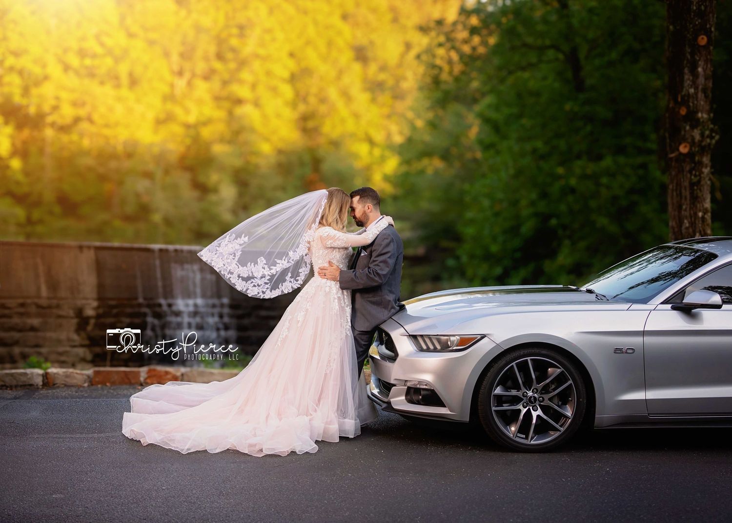A flowing white wedding dress train trails elegantly beside a sleek silver luxury car at sunset.