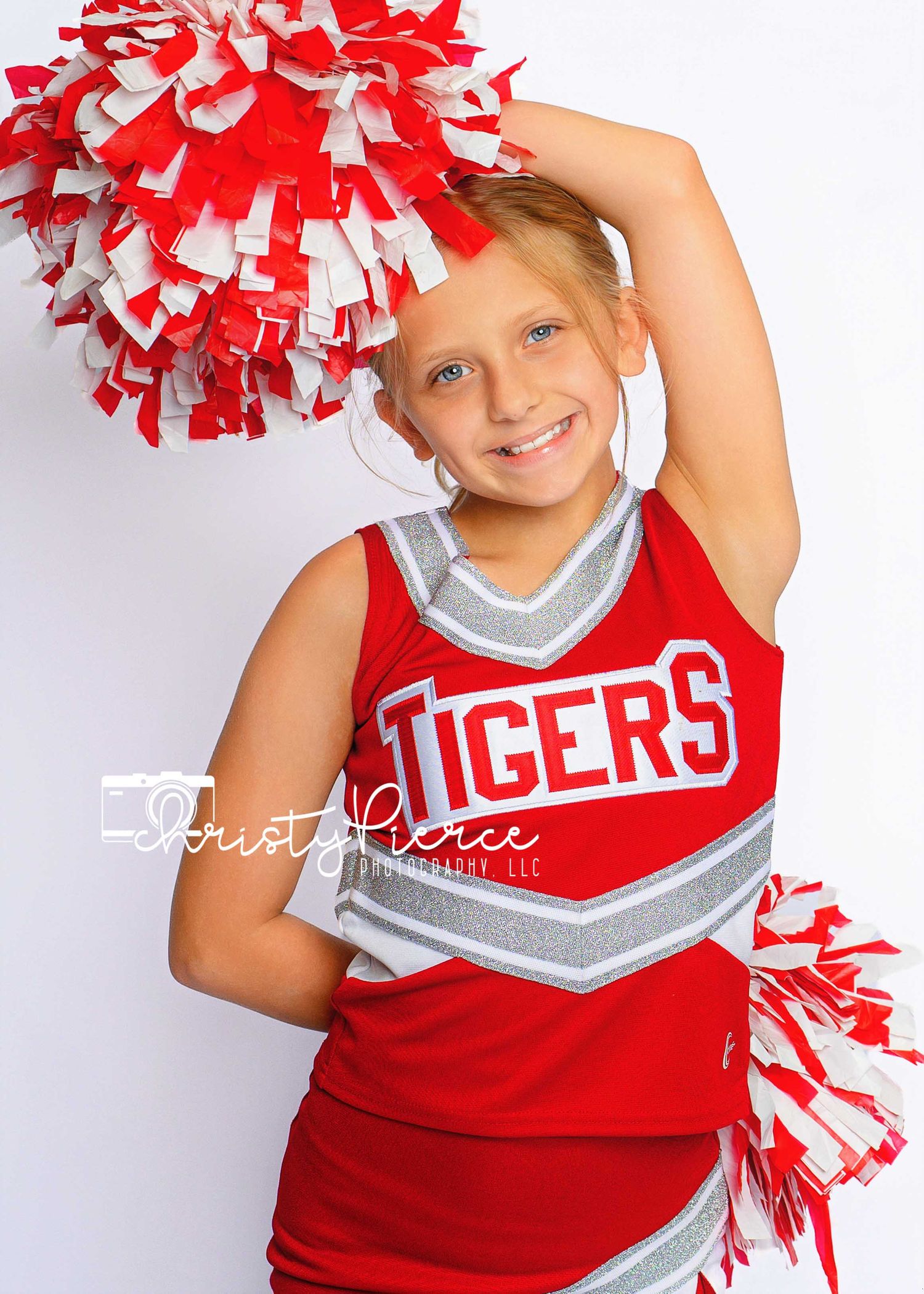 Young girl in red Tigers cheerleader uniform smiling with red and white pom-poms raised overhead.