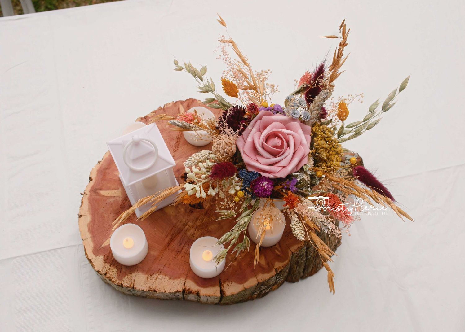 Rustic wood slice centerpiece with dried flowers, pink rose, candles, and small gift box for weddings.