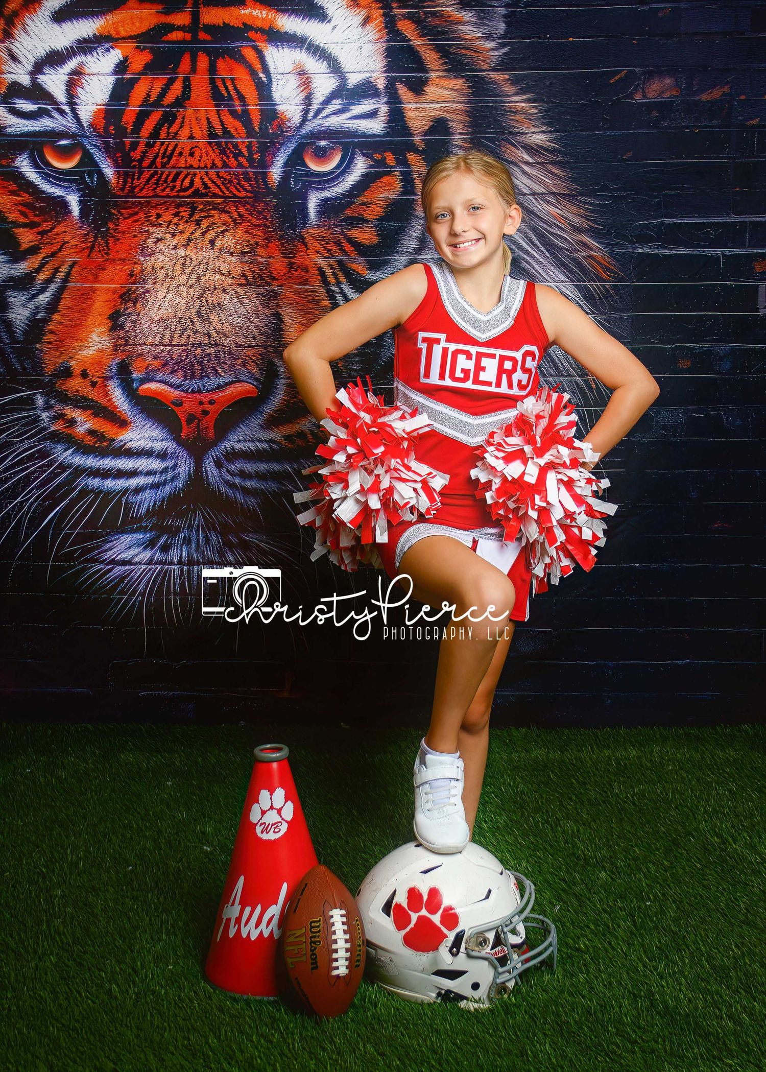 Young girl in red Tigers cheerleader uniform poses with pom-poms, football helmet, and megaphone in front of tiger mural.