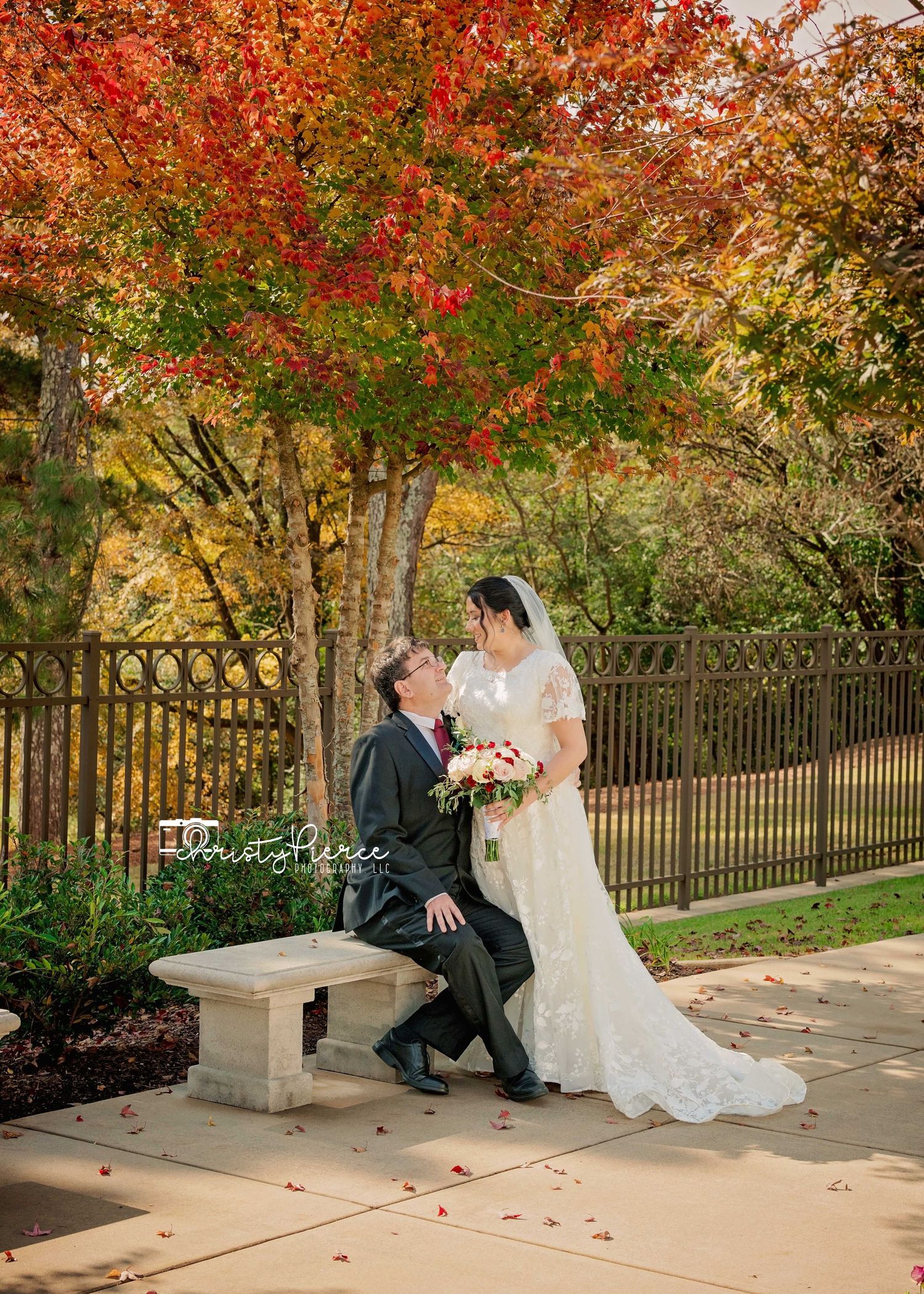 A couple sits together on a stone bench surrounded by vibrant autumn foliage in a garden setting.