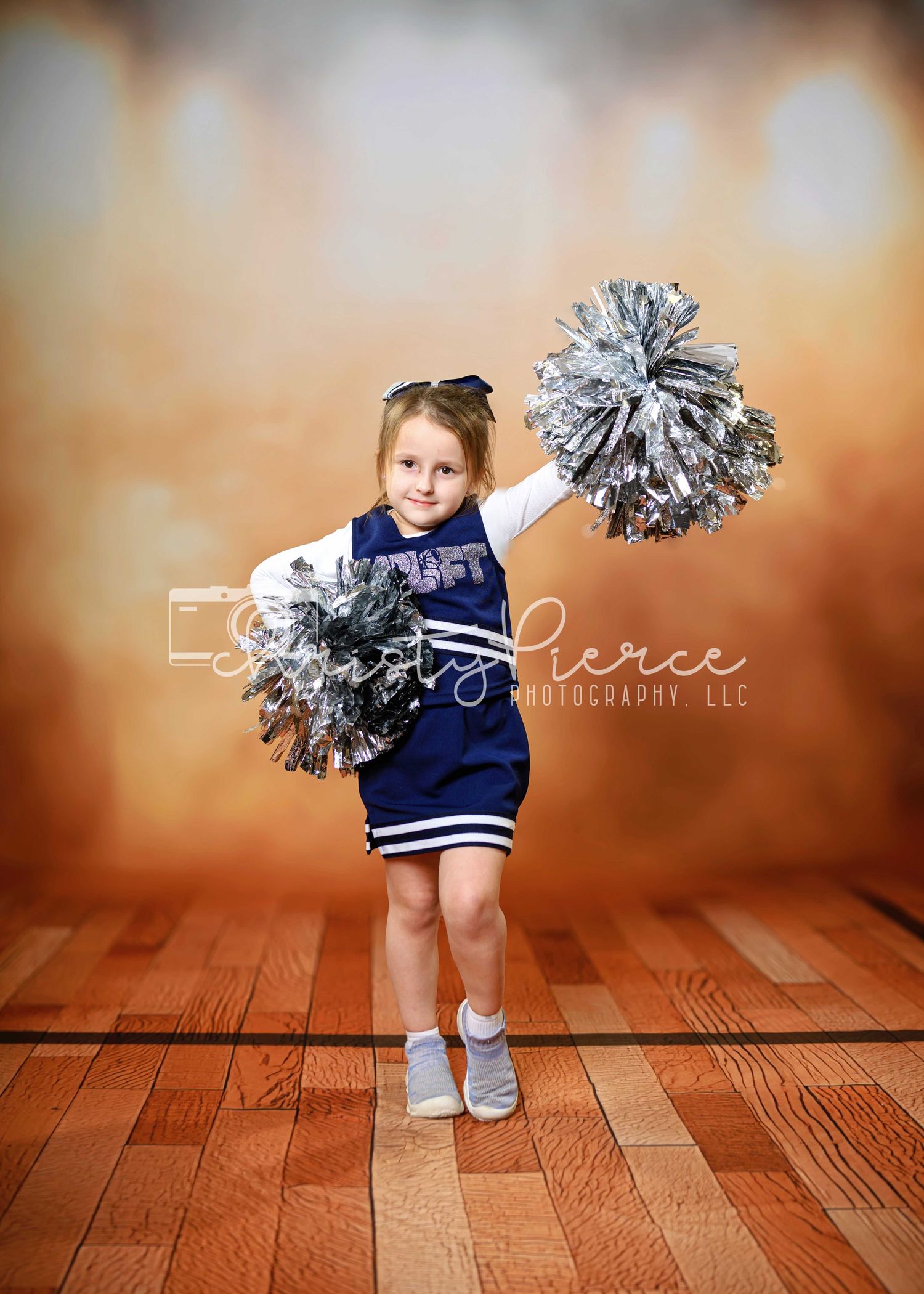 A small cheerleader in a navy uniform performs poses with silver pom poms on a wooden floor in a studio setting.