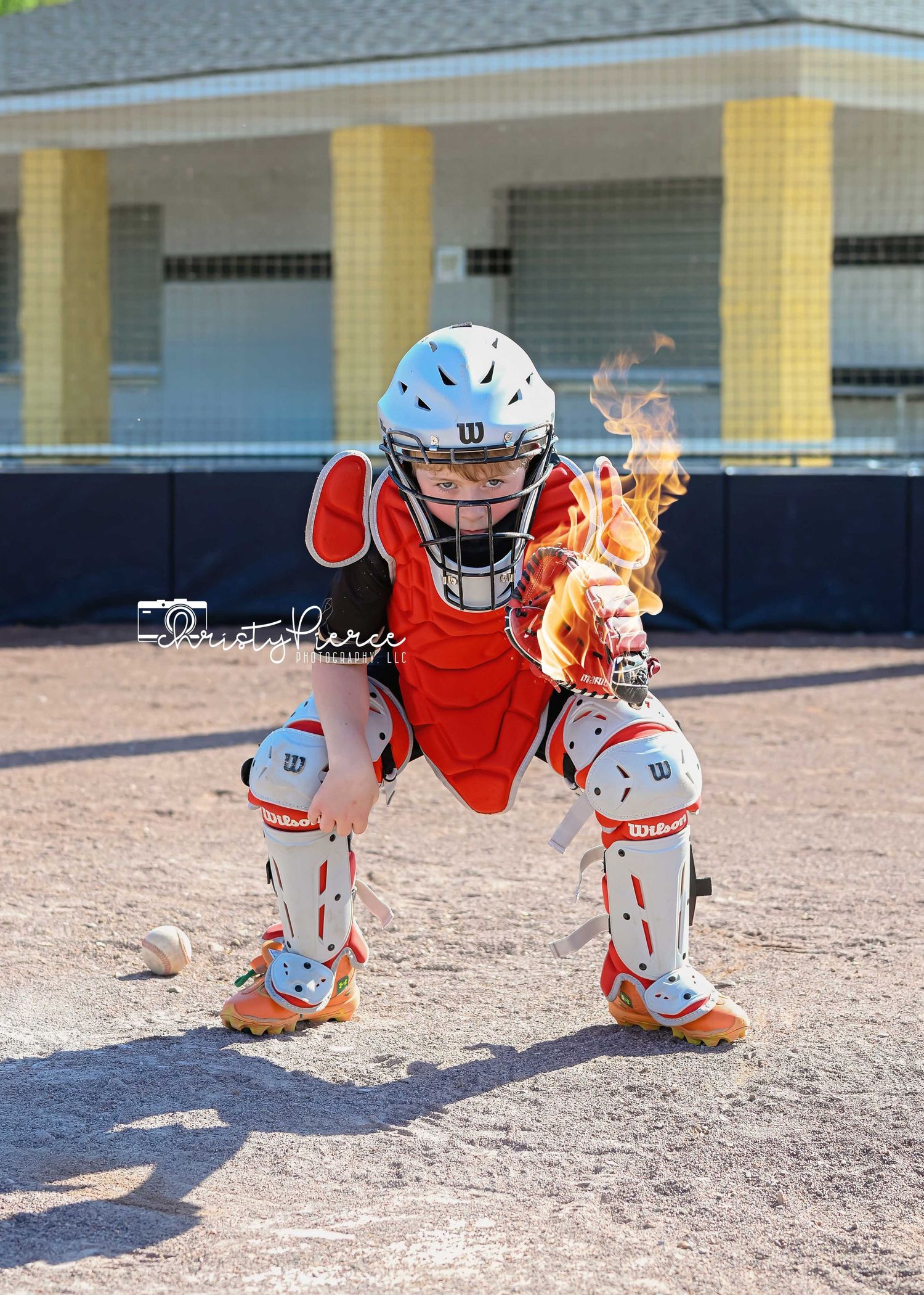 Baseball catcher in orange uniform with protective gear crouches on a dirt field as flames emerge from equipment.