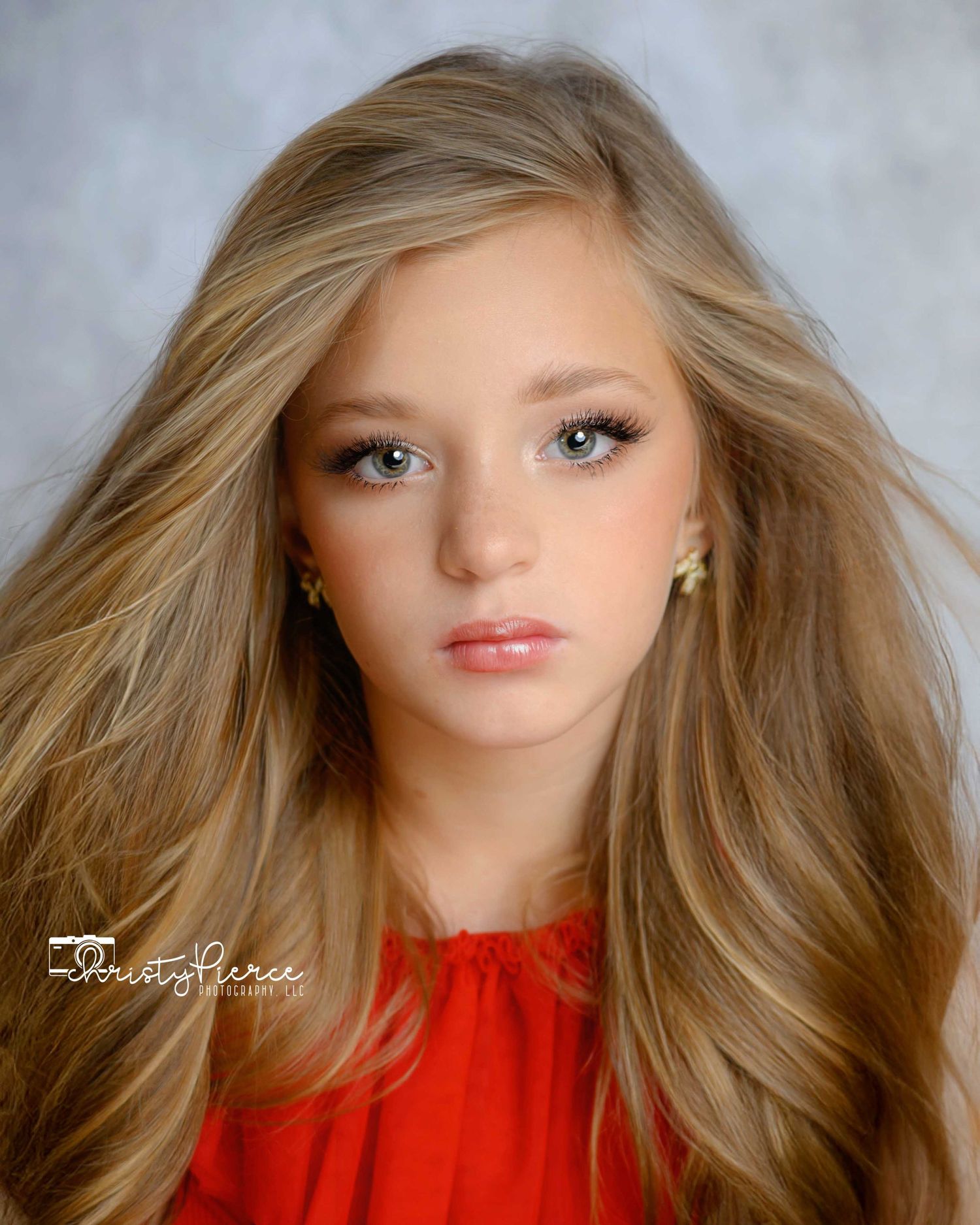 Portrait of a young person with flowing blonde hair wearing a red top against a gray studio background.