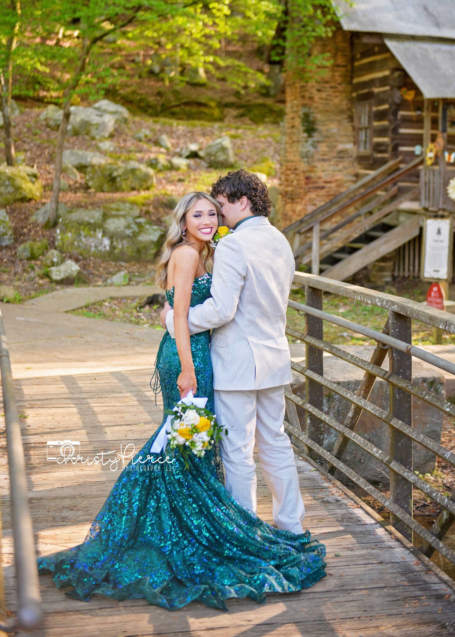 A couple in formal wear poses on a wooden bridge, with the bride wearing a stunning teal sequin gown near a rustic cabin.