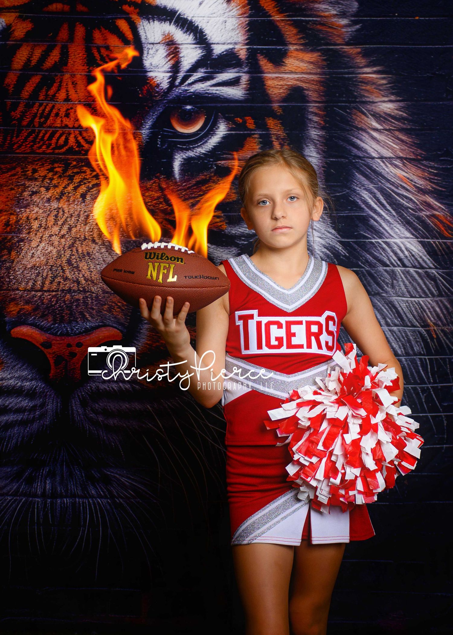 Young girl in red Tigers cheerleader uniform holds flaming NFL football, posing before a tiger mural backdrop.