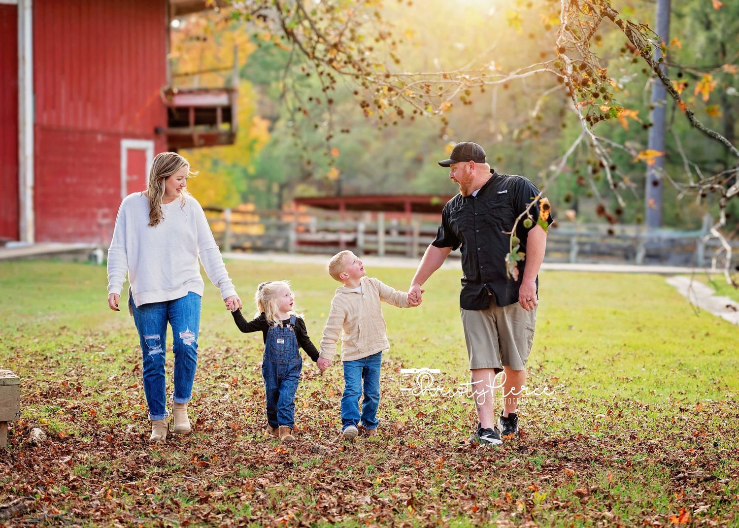 A family walks together through fallen autumn leaves near a red barn on a sunny fall afternoon.